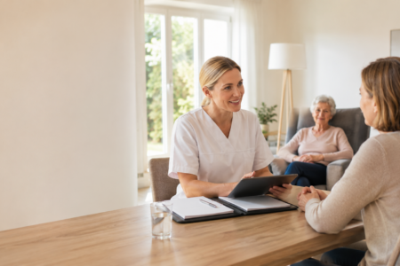 Pflegekraft berät eine Angehörige im modernen Wohnzimmer, während eine Seniorin im Hintergrund entspannt sitzt.