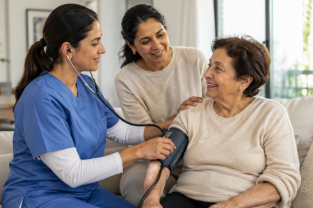 Pflegekraft misst den Blutdruck einer älteren Patientin im häuslichen Umfeld, während eine Angehörige unterstützend daneben sitzt.