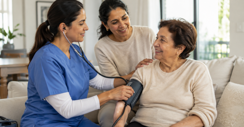 Pflegekraft misst den Blutdruck einer älteren Patientin im häuslichen Umfeld, während eine Angehörige unterstützend daneben sitzt.